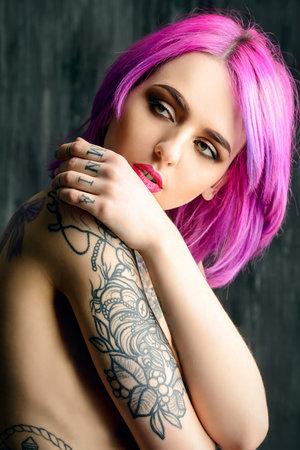 Beautiful young woman with stylish crimson hair and tattoo on her body posing over dark grunge background. Hair coloring. Cosmetics, make-up. Tattoo.の写真素材