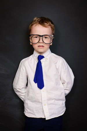 Four year old boy in white shirt and a tie posing by a school blackboard in big glasses. Educational...