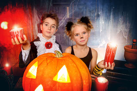 Two cute children dressed as a vampire and a black cat posing with pumpkin in a halloween room. Halloween concept.の写真素材