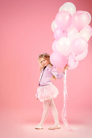 Pretty little girl with beautiful blonde hair alluring with pink balloons over pink background. Little princess with a crown on her head. Kids fashion.の写真素材