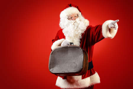 Santa Claus stands with old suitcase over red background. Christmas time.の写真素材