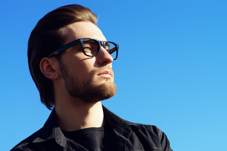 Handsome man in black jacket and spectacles posing outdoor over blue sky. Men's beauty, fashion....