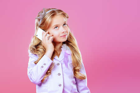 Pretty little girl with beautiful blonde hair talking on her cell phone over pink background. Little princess with a crown on her head. Kids fashion. の写真素材