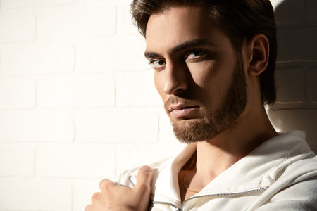 Beauty portrait of a handsome pensive young man standing by a white brick wall. Men's beauty,...