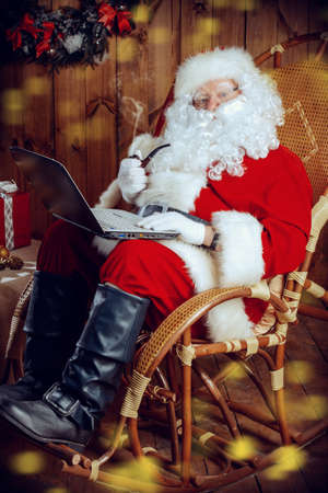 Modern Santa Claus at his wooden house working on his laptop and smoking a pipe. Christmas.の写真素材