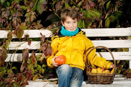 A boy sitting on a bench with a basket of red apples and pumpkin in a beautiful autumn park. Children's fashion.の写真素材