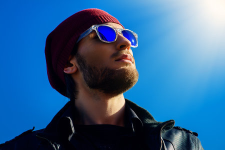 Portrait of a handsome young man in a leather jacket and sunglasses over blue sky background. Men's beauty, fashion.の写真素材