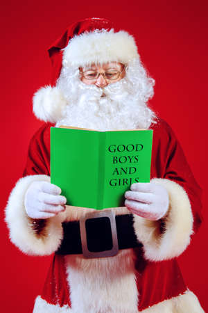 Portrait of a traditional Santa Claus over Christmas red background holding his notebook with a list of good boys and girls. Studio shot. Christmas.の写真素材