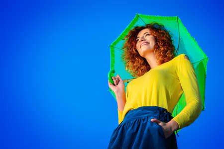 Pretty joyful young woman with beautiful foxy hair posing with umbrella over blue sky. Bright colorful clothes. Beauty, fashion. Bright lifestyle. Copy space.の写真素材