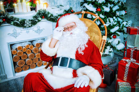 Good old Santa Claus sitting in a rocking chair in the room by the fireplace and Christmas tree, beautifully decorated for Christmas.の写真素材