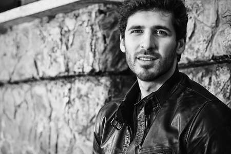 Black-and-white portrait of a handsome brunet man in black leather jacket standing by a stone wall.の写真素材