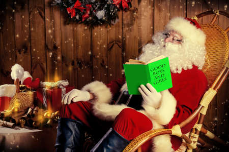 Santa Claus sitting in his wooden house in a comfortable chair and prepare gifts for Christmas. He checks his list in a notebook.の写真素材