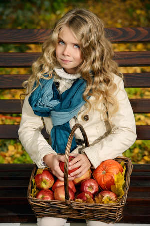 Lovely little girl sitting on a bench with a basket of red apples in a beautiful autumn park. Children's fashion.の写真素材