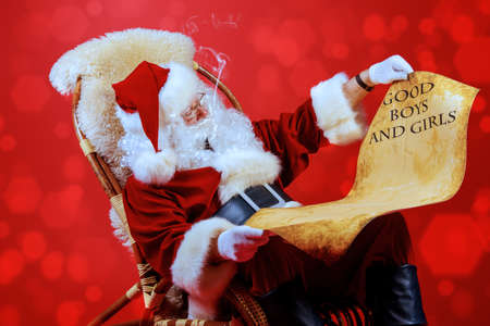 Santa Claus reading the list of presents and smoking a pipe in a rocking chair over red background. Christmas.の写真素材