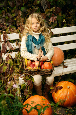 Lovely little girl sitting on a bench with a basket of red apples in a beautiful autumn park. Children's fashion.の写真素材