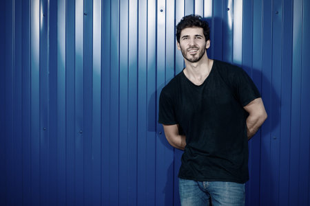 Attractive masculine man standing on a city street by a blue metal fence. Male beauty, fashion.の写真素材