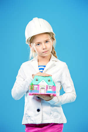 Little girl in a builder helmet  holding house in her hands. The construction industry, mortgage concept. The house for the whole family.の写真素材