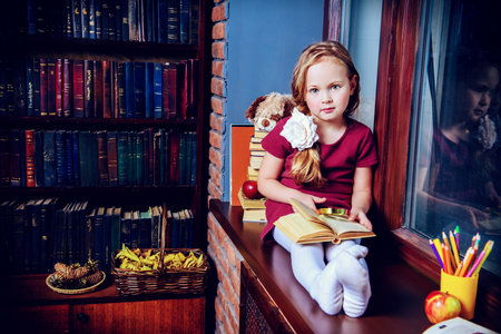 Cute little girl sits by the window with a book on her knees. Bookshelves background. Home. Education.の写真素材