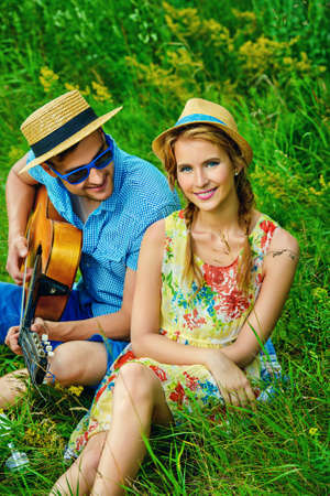 Enamored young man with a guitar singing his beloved girlfriend. They are sitting together on a lawn on a sunny summer day. Love concept.の写真素材