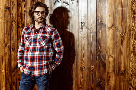 Intelligent well-groomed young man in glasses and a casual shirt posing by a wooden wall. Men's beauty, fashion. Men's barbershop, Hairstyle.の写真素材