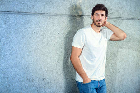 Portrait of a handsome young man in jeans and white t-shirt leaning to a concrete wall.の写真素材