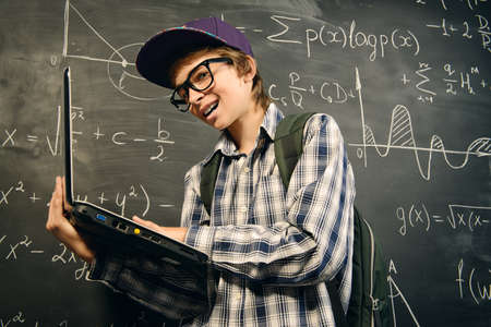 Education, high school, college. Portrait of a student boy standing by a school blackboard with his laptop.の写真素材