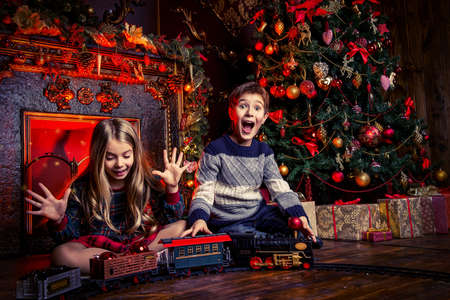 Two cheerful children playing with a toy railway near the Christmas tree and fireplace. Christmas night. Christmas decoration.の写真素材