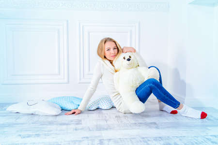 Cute teen girl in a warm knitted clothes sitting in an embrace with her teddy bear. Youth fashion, winter style. White styled interior.の写真素材