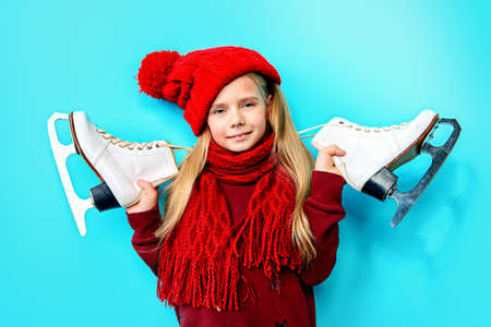 Cheerful little girl in warm sweater and hat holding figure skates. Blue background.の写真素材