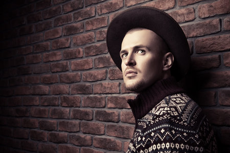 Portrait of a handsome man wearing sweater and hat standing by the brick wall. Male beauty, fashion.の写真素材