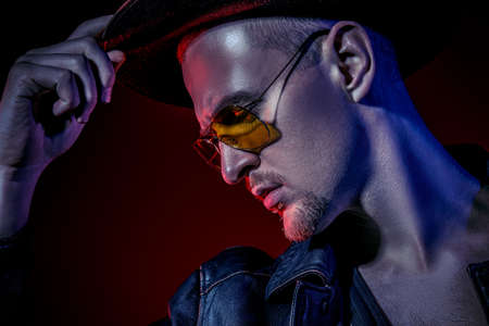 Close-up portrait of a stylish brutal man wearing leather jacket and sunglasses posing over dark red background. Male fashion.の写真素材
