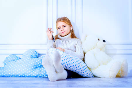 Happy 7 year old girl wearing knitted clothes sitting on a floor with her teddy bear and smart phone. Children's fashion.の写真素材