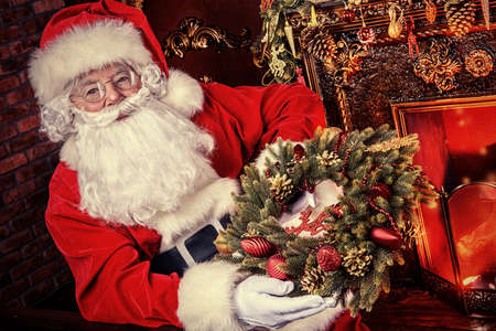 Santa Claus in his house next to the fireplace and Christmas tree holds a Christmas wreath.の写真素材