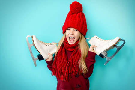 Cheerful little girl in warm sweater and hat holding figure skates. Blue background.の写真素材