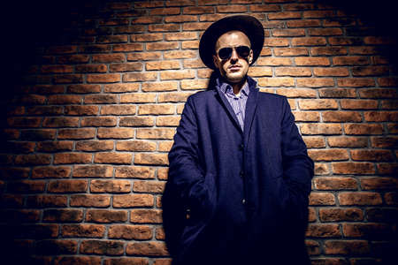 Portrait of a stylish handsome man wearing hat, coat and sunglasses. Men's beauty, seasonal fashion. Brick wall background.の写真素材