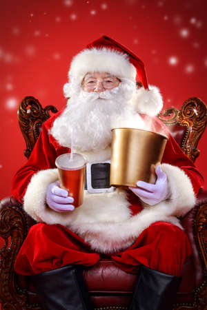 Merry Christmas and Happy New Year. Santa Claus sitting on his armchair and eating popcorn and drinking soda. Entertainment and cinema concept. Red background.の写真素材