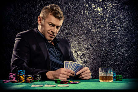 A wealthy mature man drinking brandy and playing poker with the excitement in a casino. Gambling, playing cards and roulette.の写真素材