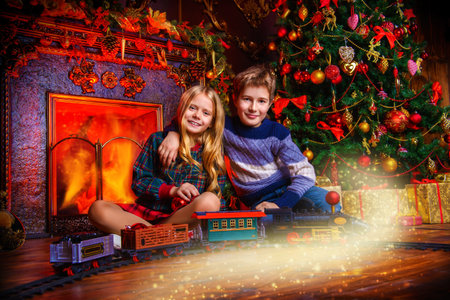 Brother and sister playing with a toy railway near the Christmas tree and fireplace. Christmas night. Christmas decoration.の写真素材