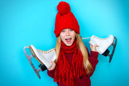 Cheerful little girl in warm sweater and hat holding figure skates. Blue background.の写真素材