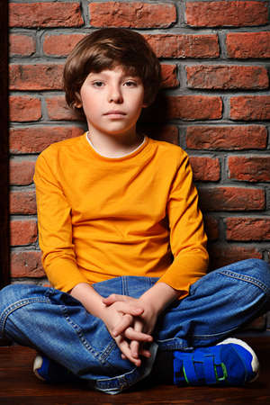 Portrait of a cute nine year old boy sitting on a floor leaning against the brick wall. Clothes for children. Kid's fashion.の写真素材