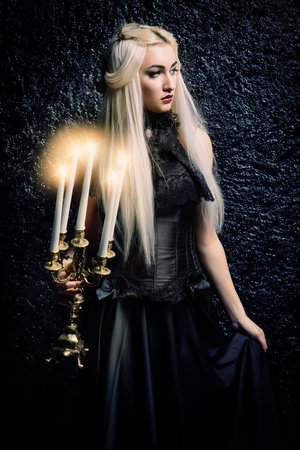 Beautiful gothic lady in long black dress standing with candles in the dark. Middle Ages. Vampires. Halloween concept.の写真素材