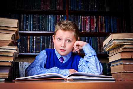 Smart boy sitting in the library by the bookshelves with many old books. Educational concept. Science.の写真素材
