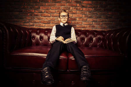 Smart boy in elegant suit and glasses sitting on a Chesterfield sofa with a book. Educational concept. Children's fashion. Vintage style.の写真素材