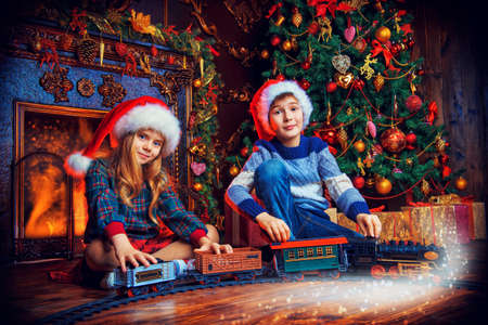 Brother and sister in Santa Claus hats playing with a toy railway near the Christmas tree and fireplace. Christmas night. Christmas decoration.の写真素材