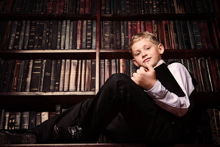 Smart boy in a suit sitting in the library by the bookshelves with many old books. Educational concept. Science.の写真素材