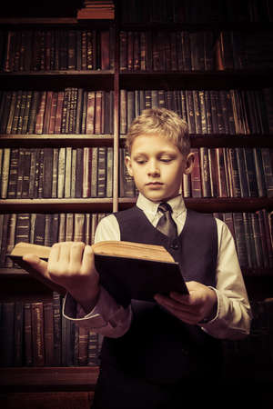 Smart boy in a suit sitting in the library by the bookshelves with many old books. Educational concept. Science.の写真素材