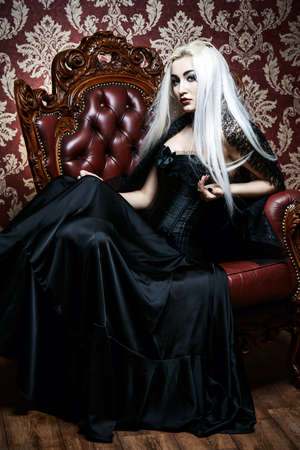 Beautiful gothic woman with long blonde hair wearing black dress. Vintage interior, castle style. Halloween.の写真素材