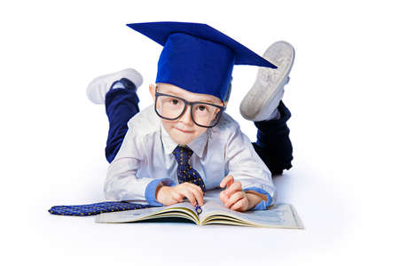 Four year old boy in white shirt, a big tie, academic hat and big glasses lying on a floor with a book. Educational concept.の写真素材