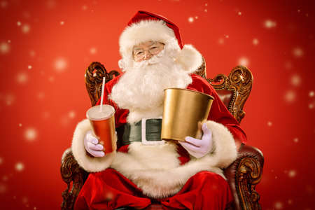 Merry Christmas and Happy New Year. Santa Claus sitting on his armchair and eating popcorn and drinking soda. Entertainment and cinema concept. Red background.の写真素材