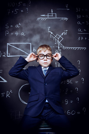 Smart schoolboy in black suit and glasses sitting on books over school blackboard.の写真素材